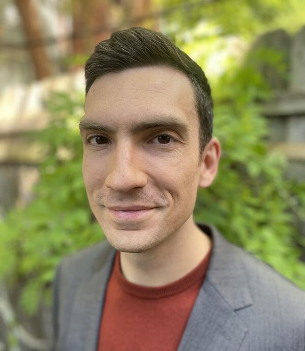 Nick Graetz poses for a headshot against a verdant backdrop. Nick has short dark hair and is wearing a gray coat and red t-shirt